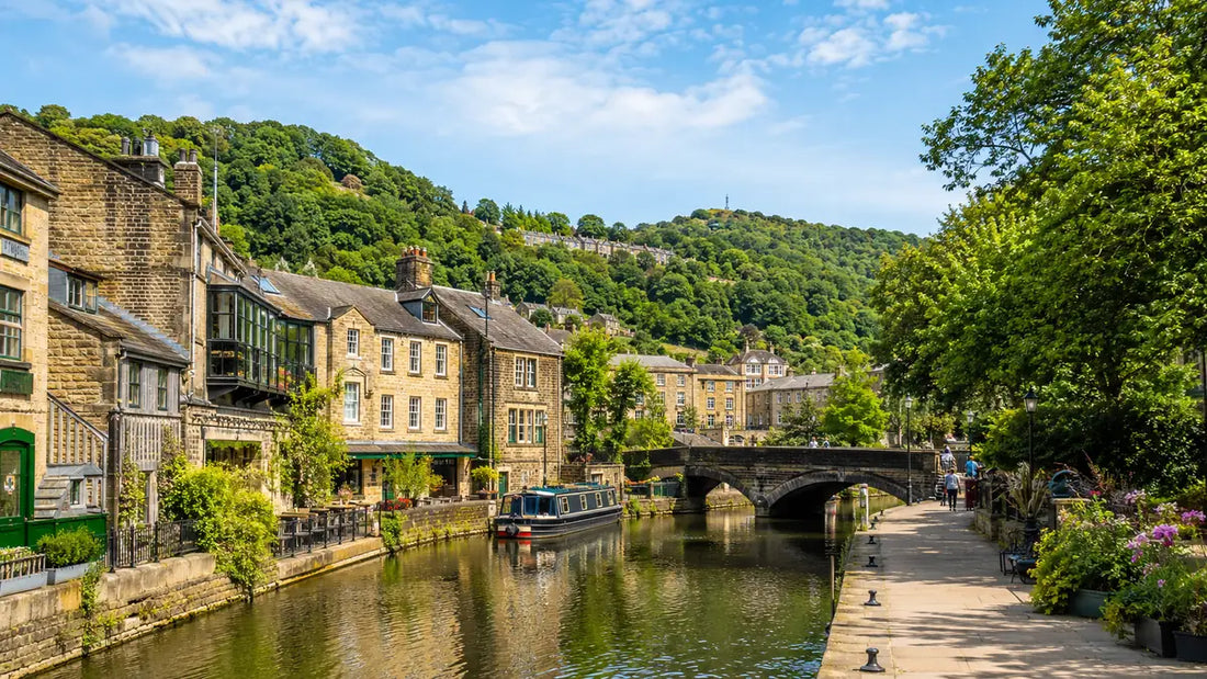 Sunny canal view in Hebden Bridge with stone buildings, a bridge, narrowboats, trees and green hills in the background.